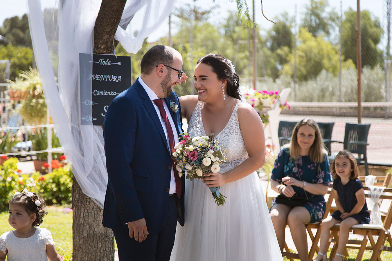 Los novios escuchando sonrientes las palabras de una invitada sentada durante su ceremonia civil al aire libre.
