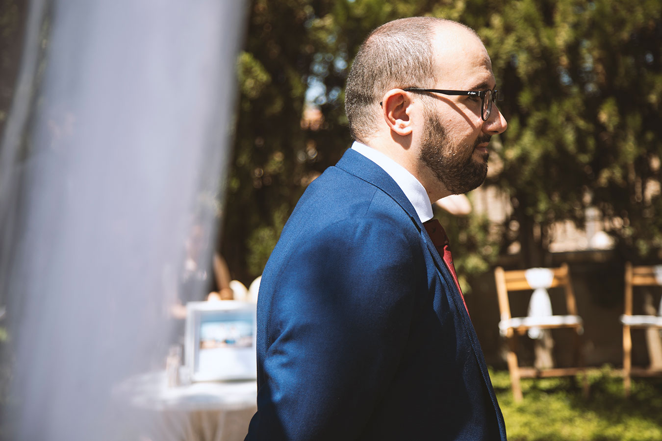 Plano medio del novio con traje azul y gafas esperando la llegada de la novia durante la ceremonia al aire libre.