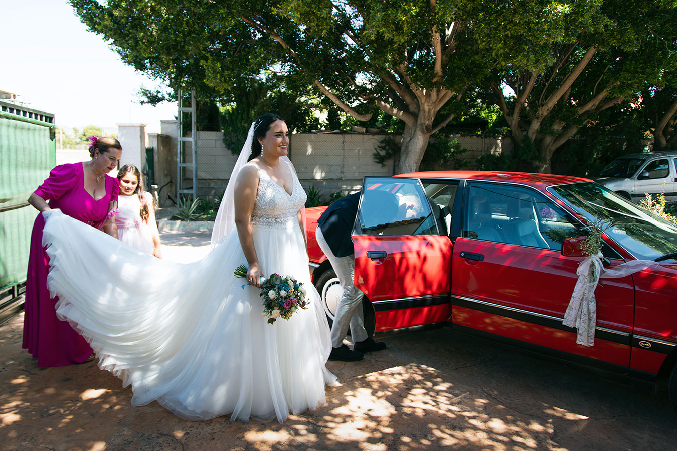 La novia luciendo su vestido con cola mientras desciende del coche clásico rojo ayudada antes de la ceremonia.