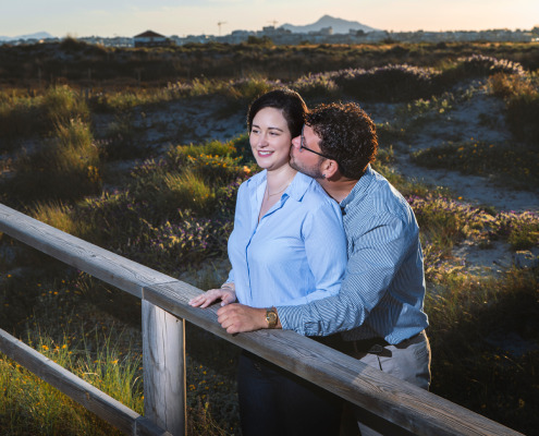Novios sonrientes apoyados en una barandilla de madera contemplando un hermoso paisaje natural en su reportaje.